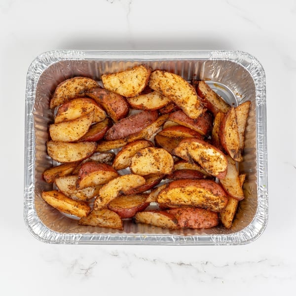 Seasoned potato wedges in a foil pan, ready for serving, on a white marble surface.