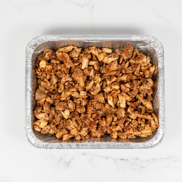 Top-down view of a foil tray filled with seasoned, diced chicken pieces on a white marble surface.