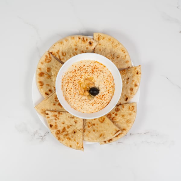 A white bowl of hummus, garnished with chickpeas, olive, and paprika, surrounded by pita bread slices.