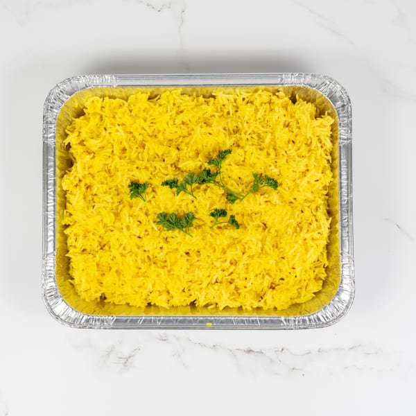Bright yellow rice with green parsley garnish in a silver foil container on a white marble surface.