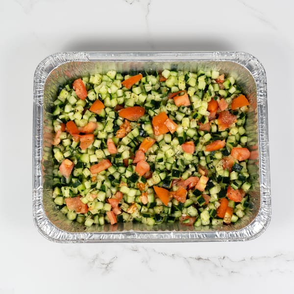 Diced cucumber and tomato salad in a silver foil catering tray on a white marble surface.