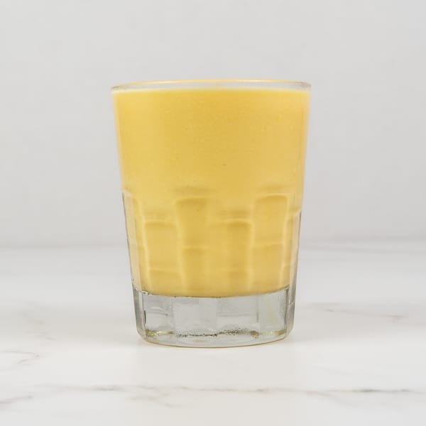 A tall glass filled with a creamy, yellow mango lassi on a white marble surface.