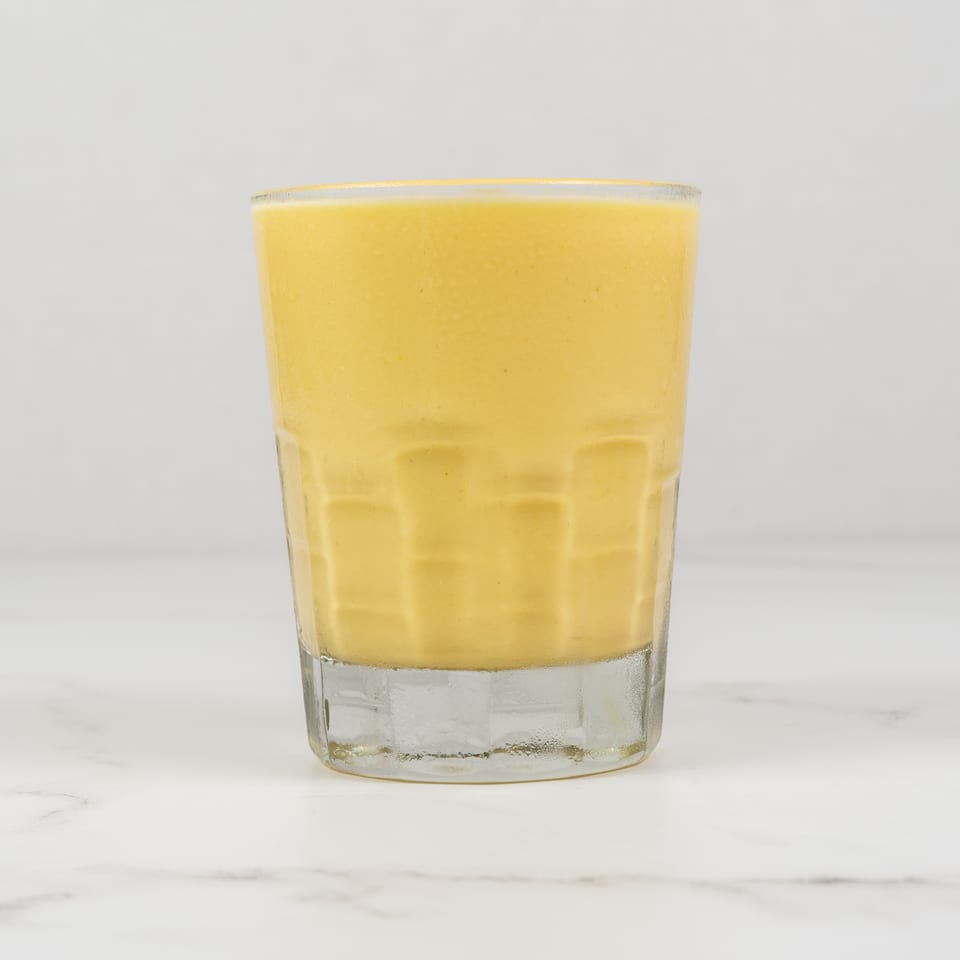 A tall glass filled with a creamy, yellow mango lassi on a white marble surface.