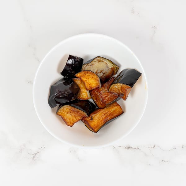 A white bowl filled with glistening, pan-fried eggplant pieces on a white marble surface.