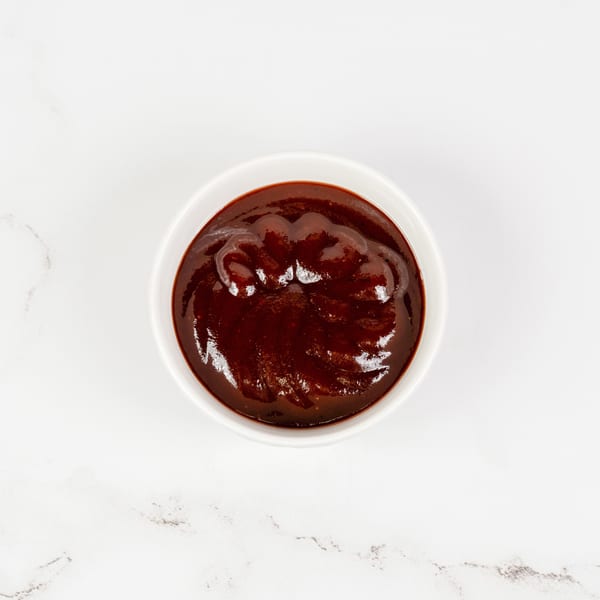 A white bowl filled with rich, dark red barbecue sauce on a white marble surface.