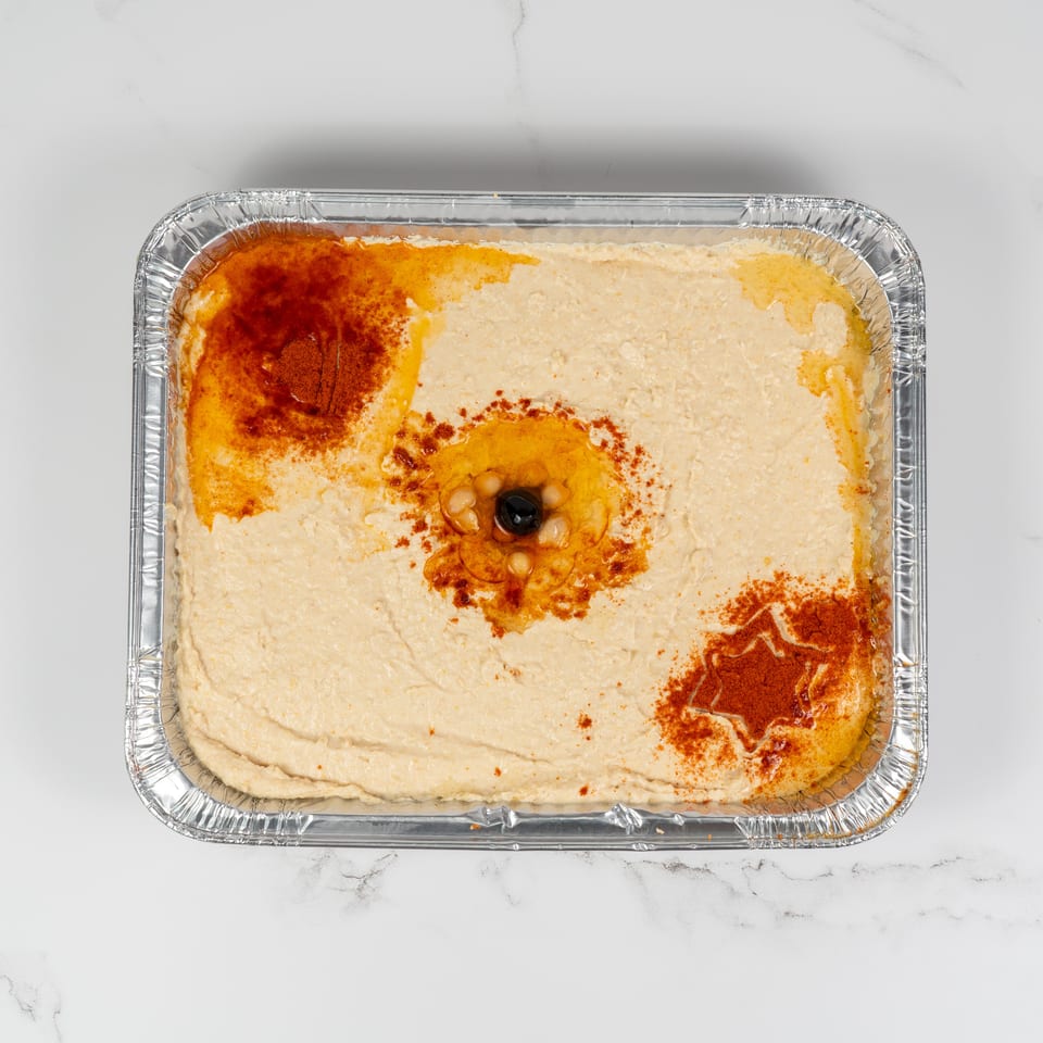 Hummus in a foil tray, garnished with paprika, olive oil, and a black olive.