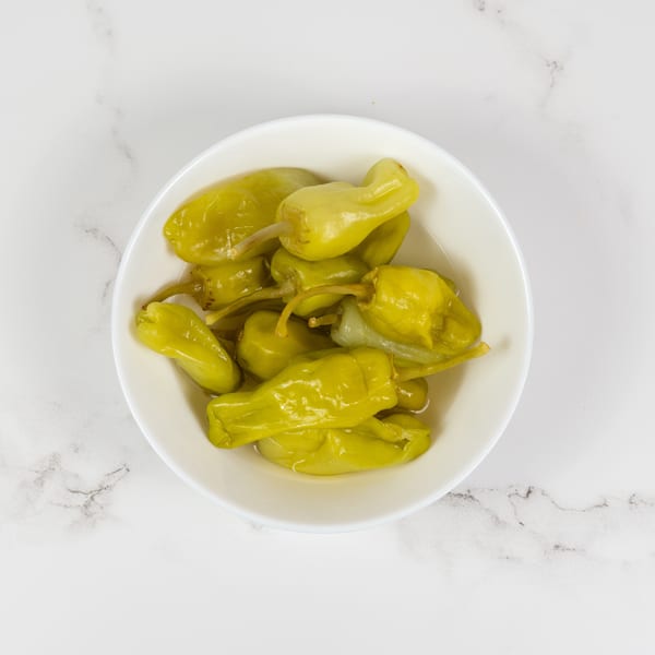 A white bowl filled with bright green pickled banana peppers, on a white marble surface.