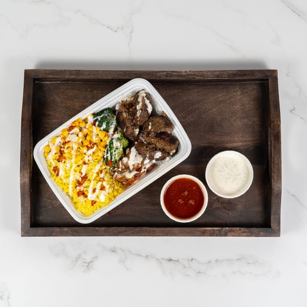 A takeout container with rice, corn, broccoli, and meat, with two sauce cups on a wooden tray.