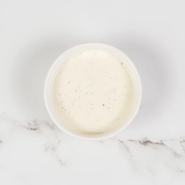 Overhead view of a small white bowl filled with creamy ranch dressing, speckled with herbs, on a marble surface.