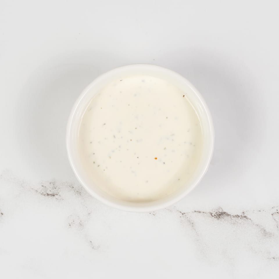 Overhead view of a small white bowl filled with creamy ranch dressing, speckled with herbs, on a marble surface.