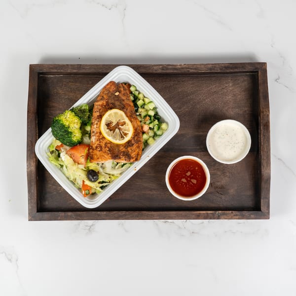Grilled salmon with lemon, broccoli, salad, and two dipping sauces on a wooden tray.