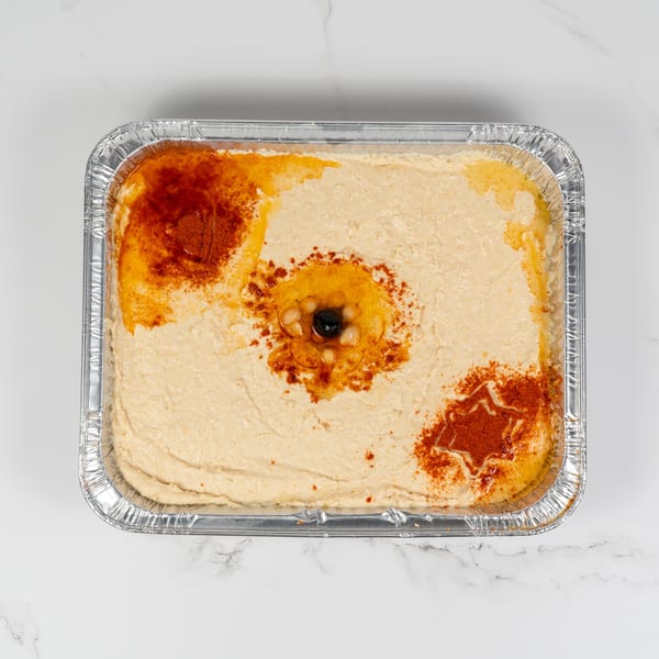 Hummus in a foil tray, garnished with paprika, olive oil, chickpeas, and a black olive.