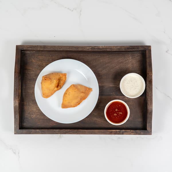 Two golden-brown samosas on a white plate, with red and white dipping sauces on a wooden tray.