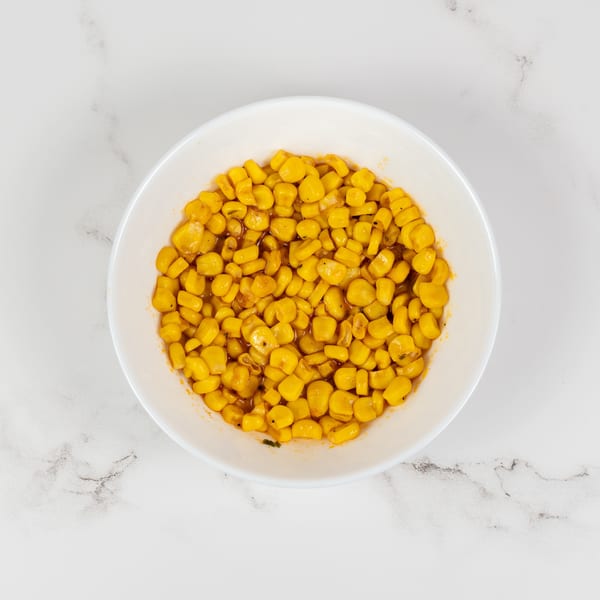 A white bowl filled with bright yellow corn kernels in a light sauce on a marble surface.