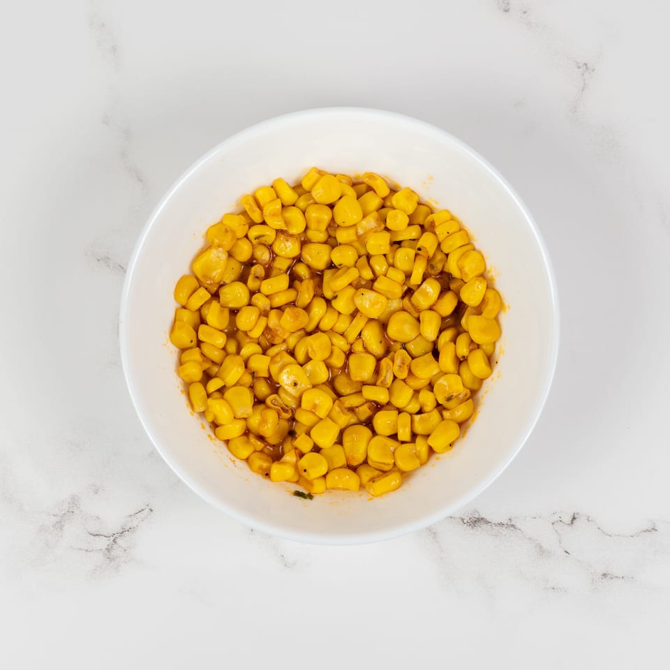 A white bowl filled with bright yellow corn kernels in a light sauce on a marble surface.